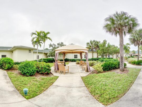 Elderly woman in a wheelchair being gently guided by a caregiver through a sunlit sensory garden in Fort Myers. Surrounded by vibrant flowers, greenery, and a winding path, the scene highlights the calming and therapeutic benefits of outdoor memory care spaces.